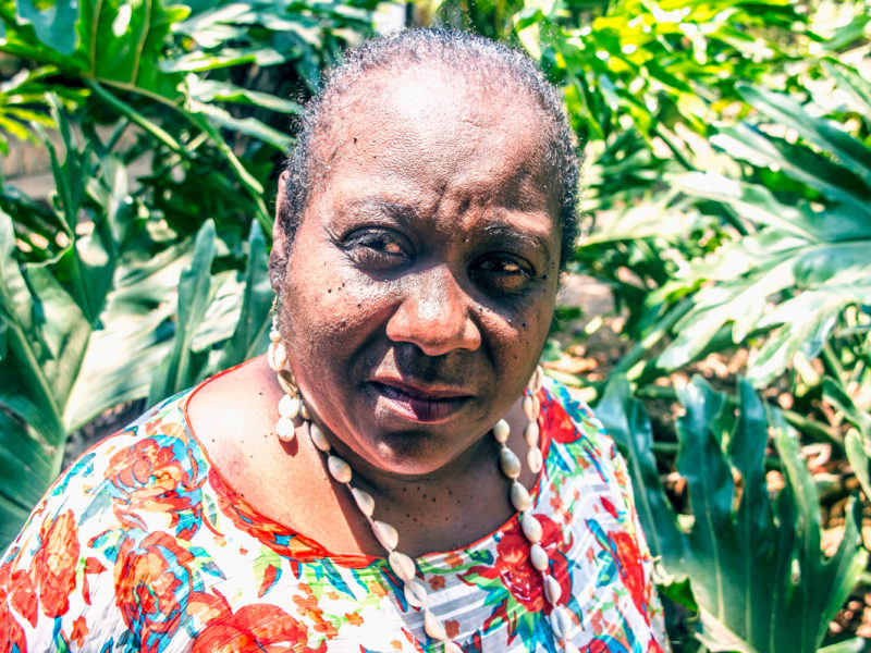 Antonieta veste uma camisa florida e olha para a câmera. Ao fundo, plantas verdes