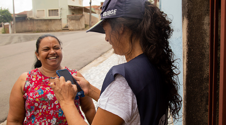 #PraCegoVer À direita, recenseadora do IBGE com dispositivo móvel de coleta e à esquerda, uma senhora respondendo ao questionário