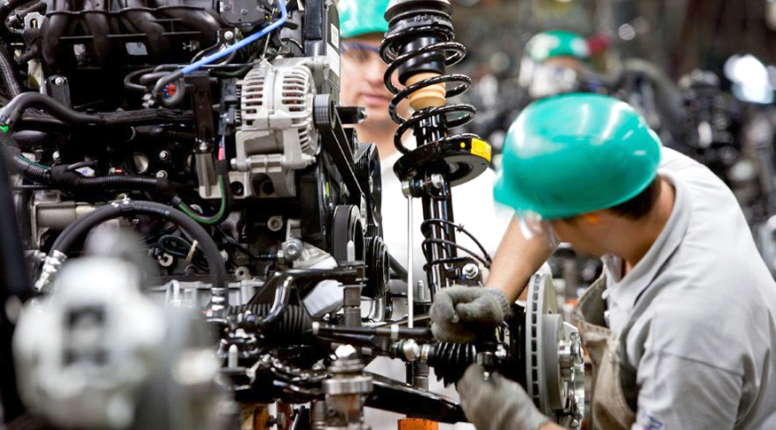 #PraCegoVer A foto mostra no primeiro plano um operário de capacete e luvas operando uma máquina industrial