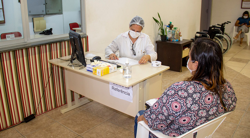 #PraCegoVer A foto mostra uma sala de atendimento médico, em que a profissional de saúde está vestida de branco e usando EPIs e a mulher que está sendo atendida, está usando uma máscara branca. Ambas estão sentadas.