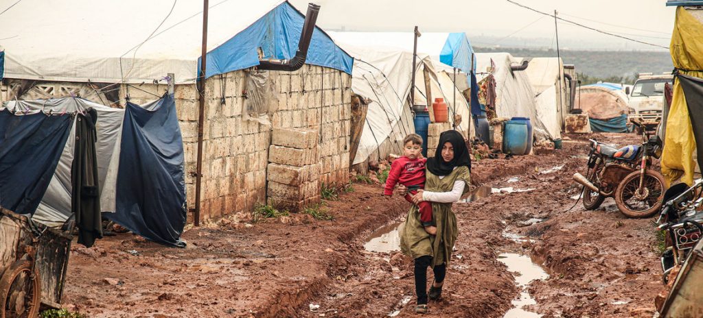 Menina caminha pela lama carregando seu irmão mais novo no campo de deslocados internos Khair Al-Sham em Idlib, na Síria. Foto: UNOCHA