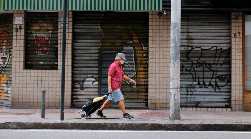 #PraCegoVer A foto mostra um homem usando máscara, empurrando um carrinho de compras, andando por uma rua vazia, com as lojas fechadas.