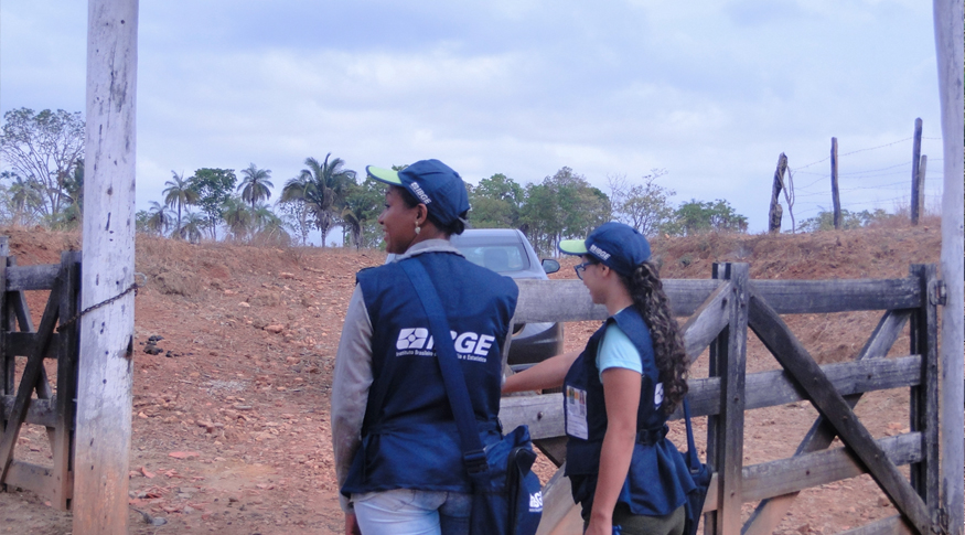 #PraCegoVer Duas recenceadoras com uniforme do iBGE na porteira de uma fazenda