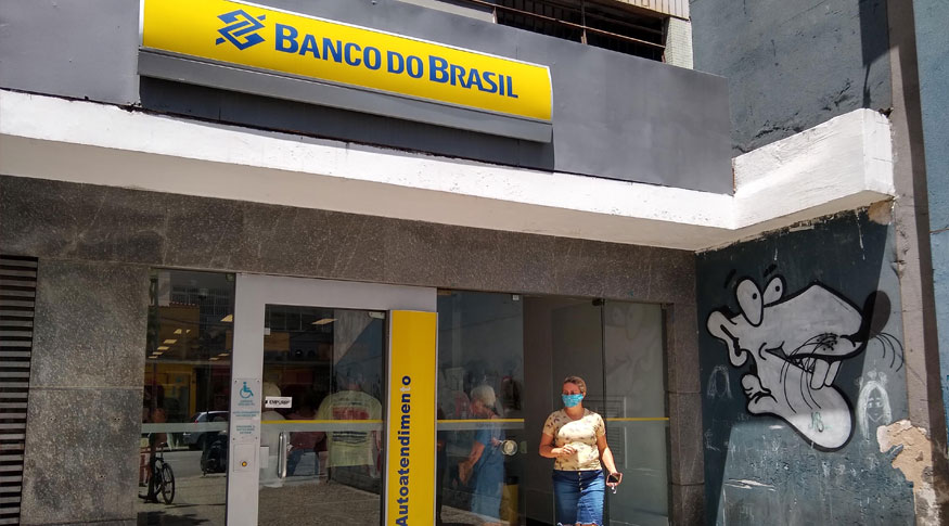 #PracegoVer Fachada do Banco do Brasil com uma mulher usando saia jeans e blusa rosa e máscara saindo da agencia. Na parede ao lado direito um grafite de um rato