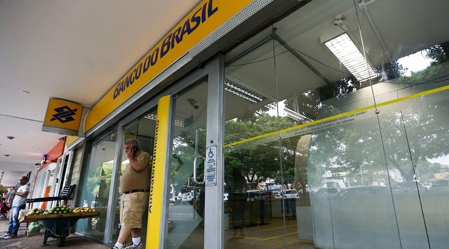 #PraCegoVer fachada de uma agencia do Banco do Brasil com um homem de blusa branca saindo da agÊncia falando ao telefone.