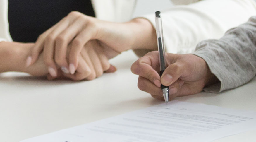 #PraCegoVer Uma mão assinando um documento em priemiro plano e ao fundo duas mãos de mulher com unhas pintadas de cor clara, cruzadas e apoiadas na mesma mesa 