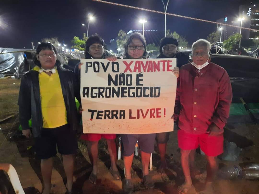 Indígenas Xavantes seguram cartaz escrito "Povo Xavante não é agronegócio. Terra Livre" durante acampamento em Brasília contra o PL 490 e a tese do Marco Temporal.