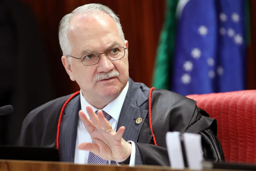 Ministro Edson Fachin, homem branco, com cabelo e bigode grisalhos, veste roupa preta e óculos redondos. Ao fundo está a bandeira do Brasil.