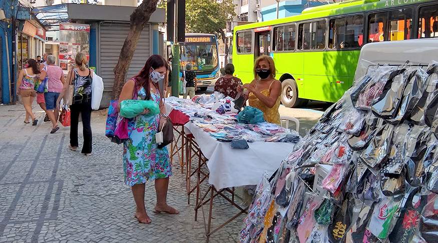 #Pracegover A foto mostra um trecho de uma rua de centro urbano. Ao fundo do lado esquerdo, aparecem pessoas caminhando. No centro, uma mulher, de vestido estampado, carregando um embrulho azul está parada em frente a uma barraca que vende máscaras de tecido. A vendedora também está de máscara e aparece do lado direito da foto, atrás da barraca. Na lateral direita, aparecem ônibus urbanos, o mais destacado é na cor verde.