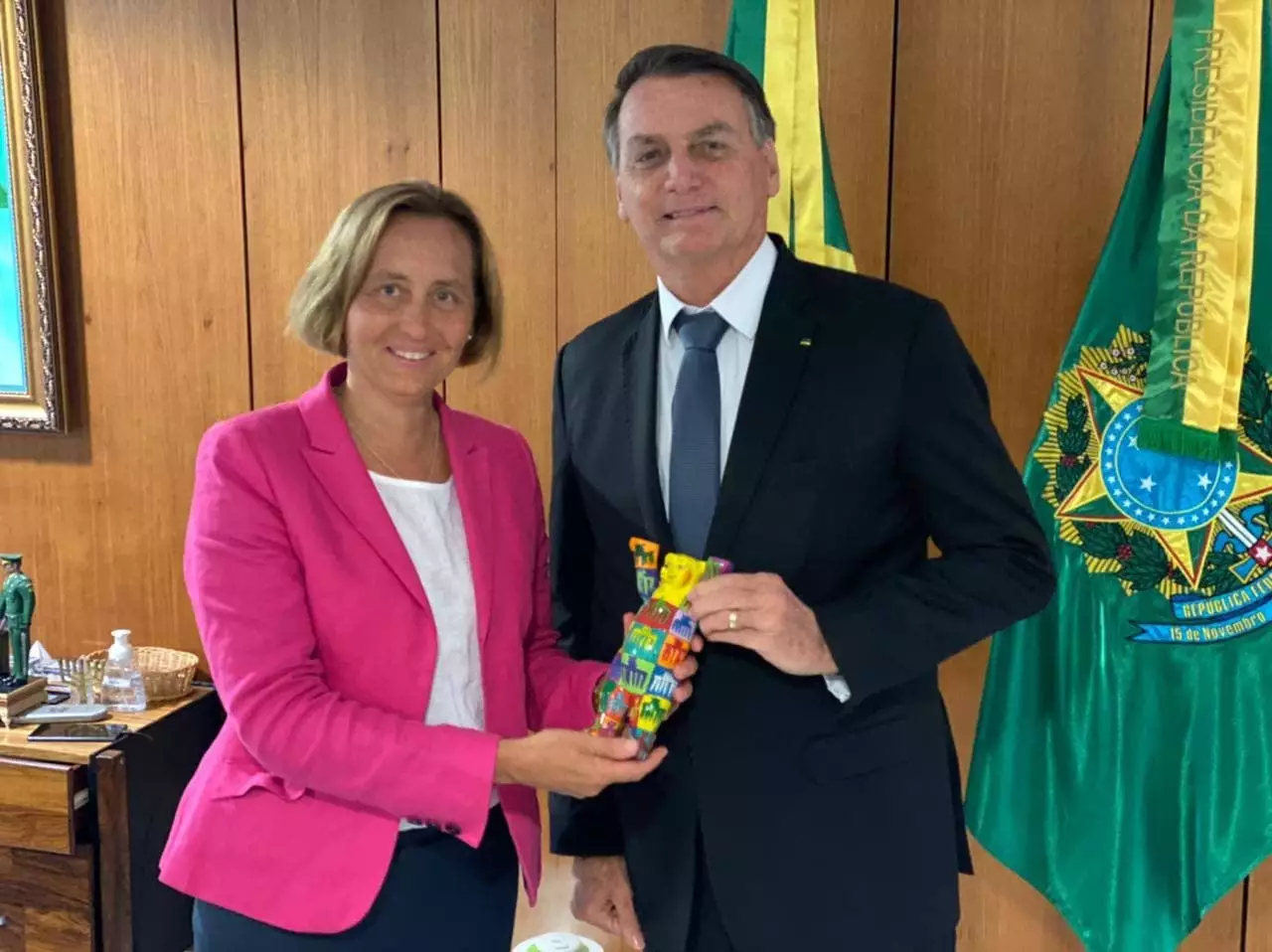 À esquerda está Beatrix von Storch, mulher branca de cabelo loiro curto e blazer rosa. À sua direita está Jair Bolsonaro, homem branco de cabelo escuro e blazer preto com gravata azul.