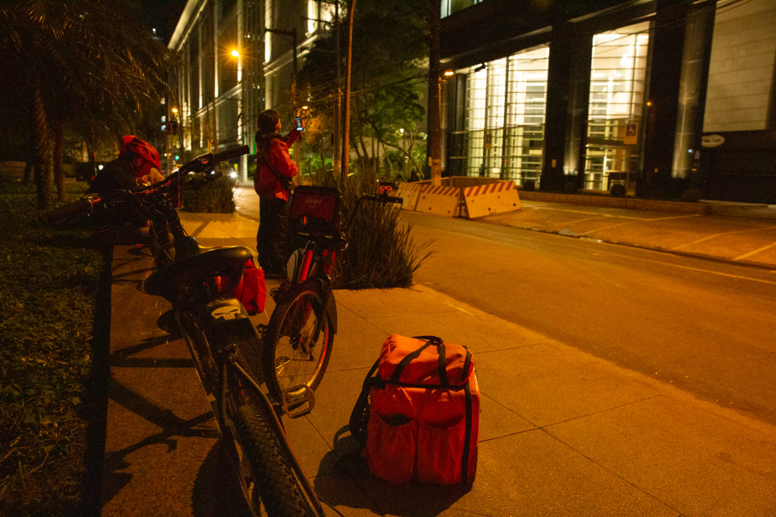 Parada na calçada, está um jovem entregador buscando sinal de telefone, com o aparelho levantado em direção ao céu; ao seu lado está algumas bicicletas e mochilas térmicas