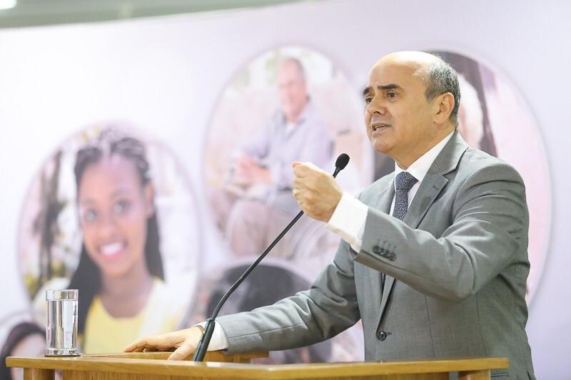 Luciano Mariz é um homem branco na faixa dos 50 anos, com cabelo calvo e grisalho; ele veste um terno cinza e está em pé em uma mesa com microfone