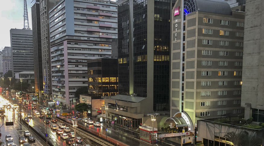 #PraCegoVer Vista aérea da cidade de SãoPaulo com a Avenida Paulista no canto inferior esquerdo da foto e 
 o hotel IBIS do lado dirito