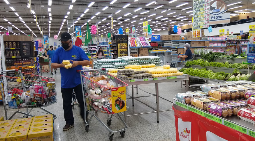 #PraCegoVer Homem de blusa azul segurando uma caixa de ovos num corredor de supermercao. A frente tem um carrinho de ocmpras com algumas compras e por traz uma pilha de caixa de ovos. O correr de lampadas em perspectiva no teto ampliam a profundidade na imagem