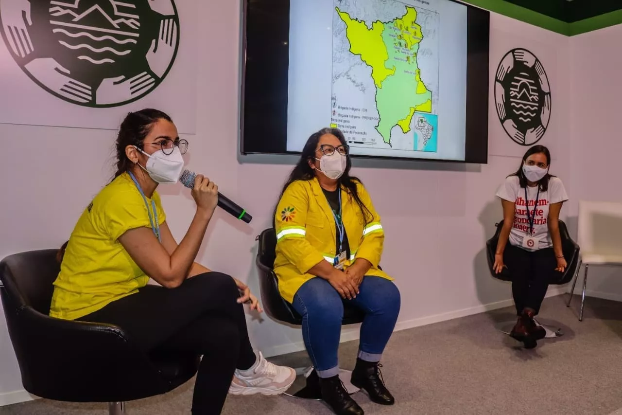 Vestida com um casaco amarelo, jeans azul e sapato preto, Sineia do Vale está ao centro entre duas outras mulheres durante painel indígena na COP-26