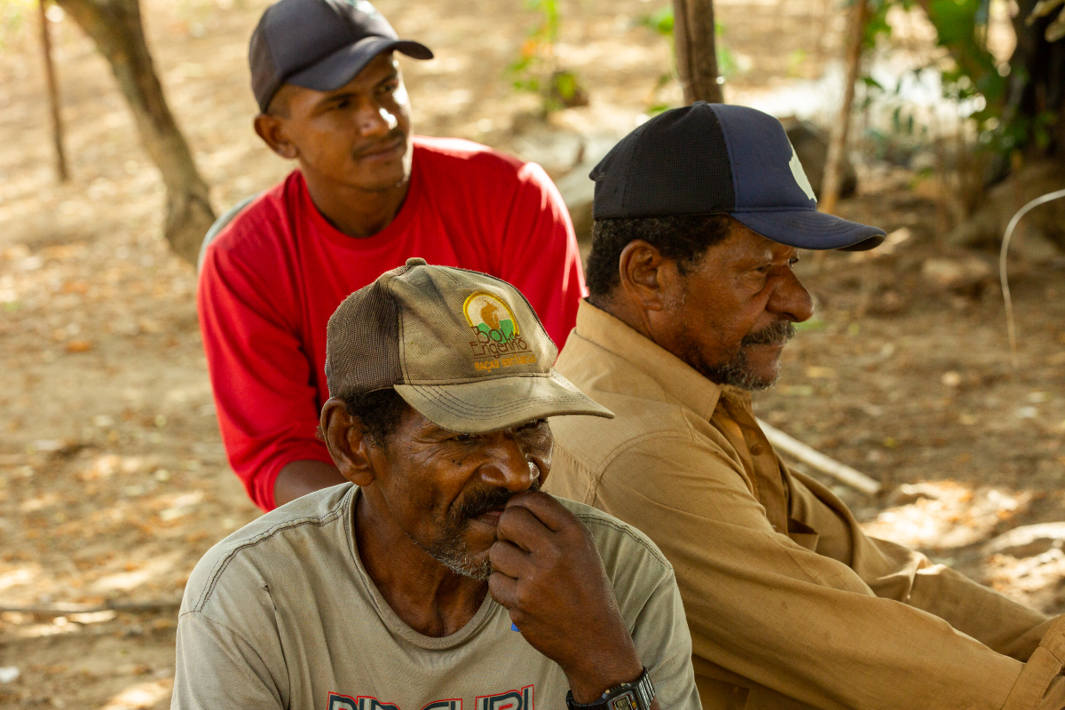 A imagem mostra três homens negros, dois mais adultos e um jovem; o primeiro veste uma camiseta vermelha e um boné cinza escuro, o segundo veste uma camisa marrom com um boné preto e azul e o terceiro veste uma camiseta cinza com um boné cinza