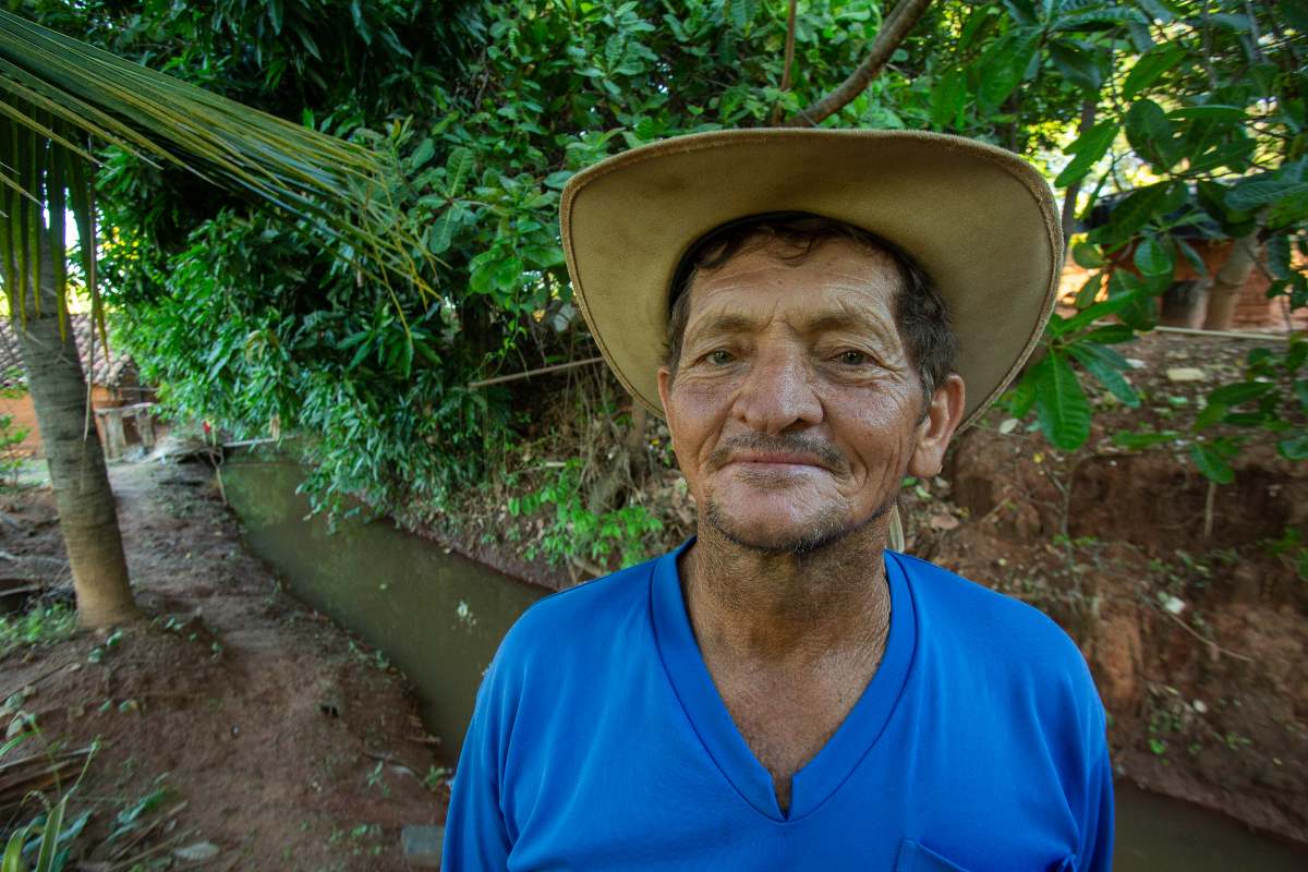 Na foto, um homem negro veste um chapéu de palha marrom com uma camiseta azul; ele aparece em frente à uma área verde com árvores e plantas rasteiras