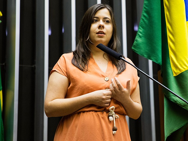 Sâmia é uma mulher branca com cabelos médios e castanhos, ela veste um vestido laranja claro