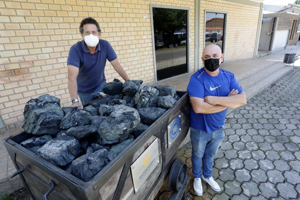 Genoir e Leonor são dois homens brancos; eles vestem camisetas na cor azul e posam ao lado de um carrinho com carvão