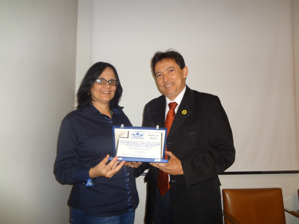 Damares Alves é uma mulher branca com cabelos escuros, ela veste uma camisa azul e óculos; ao seu lado está um homem com terno preto, com o qual ela segura um certificado