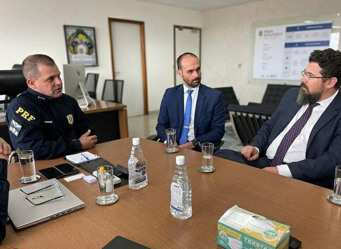 Sentado entre dois homens, está Eduardo Bolsonaro, deputado e filho do presidente; Eduardo é um homem branco, com cabelo e barca castanhos, ele veste um terno azul