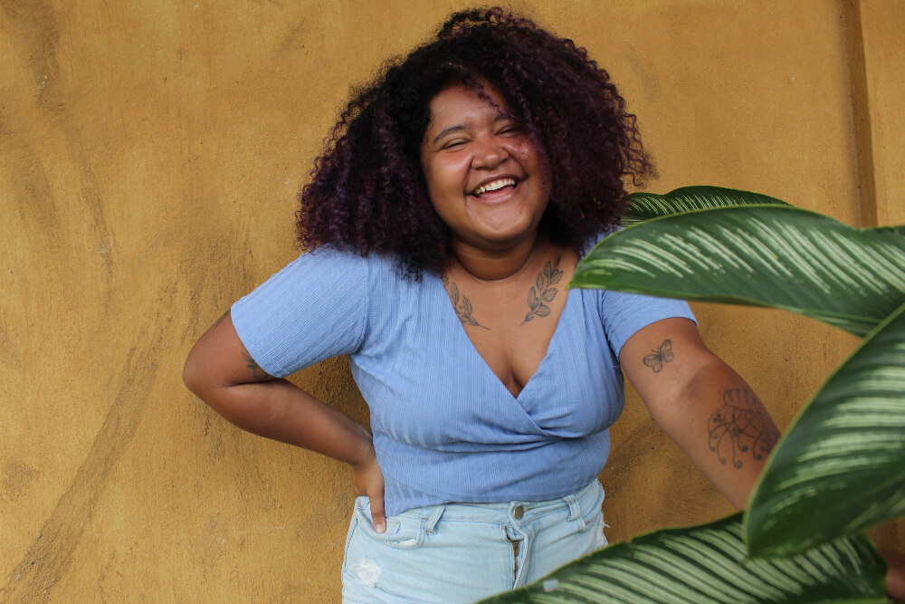 Ana Rosa é uma jovem negra com cabelo castanho cacheado; ela veste uma camiseta azul clara e está sorrindo para a foto