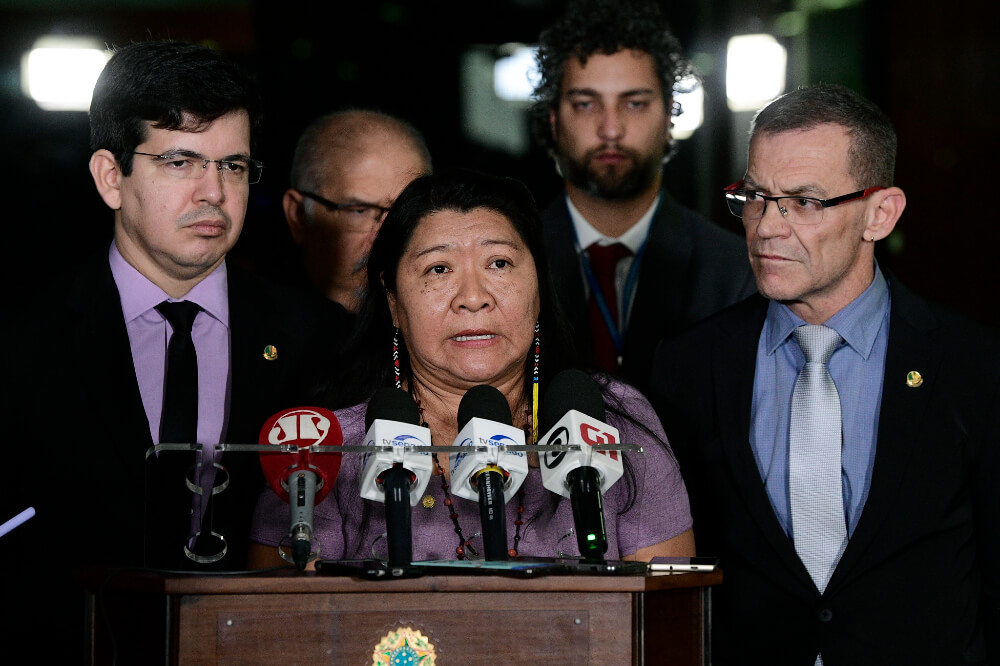 Joênia é uma mulher indígena com cabelos castanhos; na imagem ela está em um pequeno palanque com microfones de diversas emissoras, atrás dela há alguns assessores