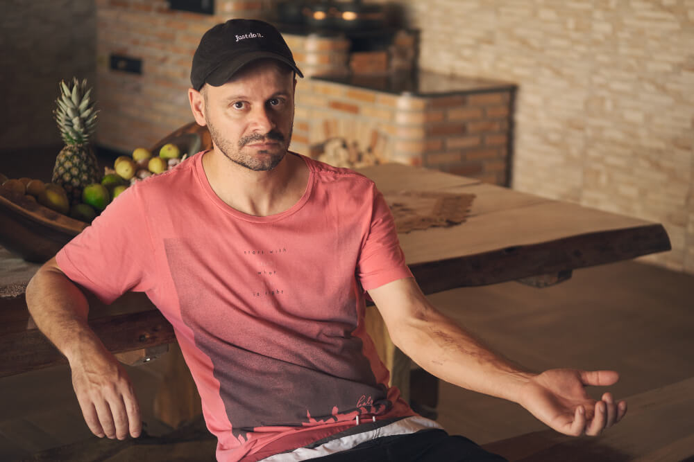 Leandro é um homem branco, com olhos e barba castanha; ele veste um boné preto e uma camiseta rosa. Sentado em uma mesa, ele está com o braço esquerdo esticado, mostrando uma cicatriz decorrente da tragédia de Brumadinho