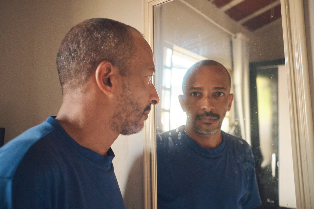 Edivaldo é um homem negro com cabelos grisalhos ralos; ele veste uma camiseta azul e está olhando para a câmera através do seu reflexo no espelho