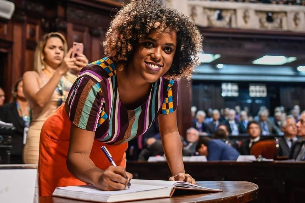 Dani Monteiro segura uma caneta enquanto assina um documento e sorri para a câmera, ao seu redor estão deputados e deputadas da Alerj