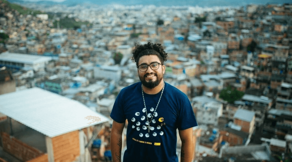 Raul Santiago é um homem negro com barba, cabelo e olhos castanhos; ele veste uma camiseta azul escuro e óculos quadrados. Atrás dele está uma favela, desfocada pela câmera