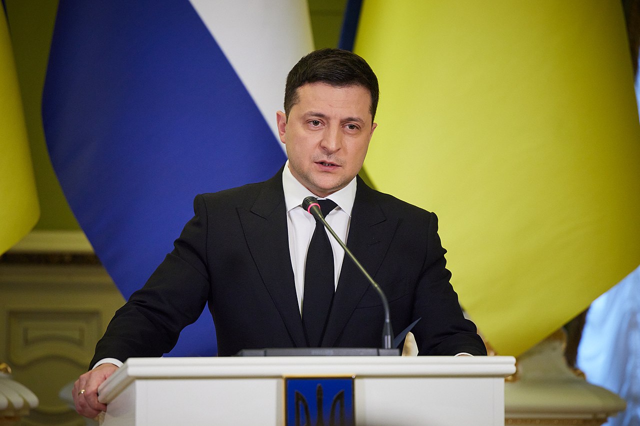 Zelensky é um homem branco na faixa dos 40 anos, tem olhos e cabelos castanhos, ele veste um terno preto com gravata preta; na imagem Zelensky está em cima de uma palanque discursando