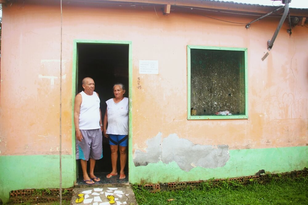 Casal de senhores posa em frente a porta de entrada de sua residência