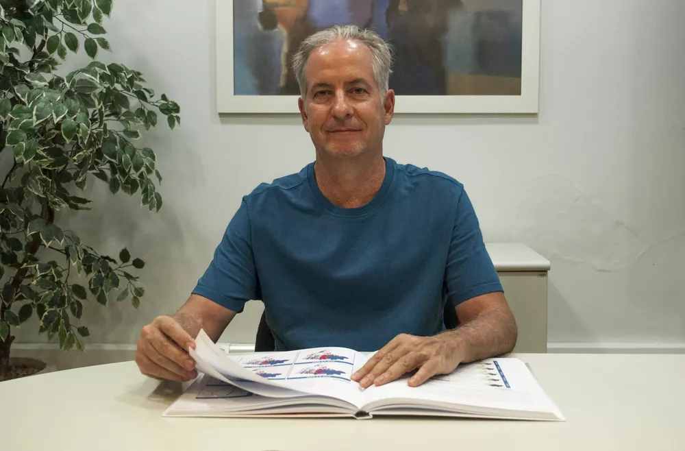 Roberto é um homem branco na faixa dos 60 anos, com cabelos grisalhos e olhos castanhos; ele veste uma camiseta azul, está sentado em uma mesa branca segurando um relatório nas mãos