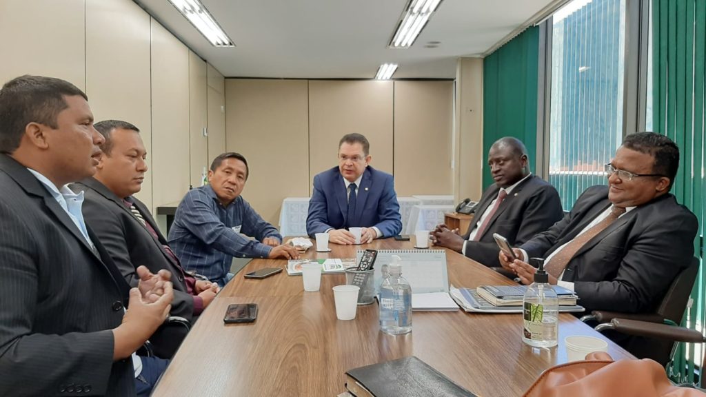 Na imagem é possível ver seis homens de terno e roupa social reunidos em uma mesa retangular de madeira