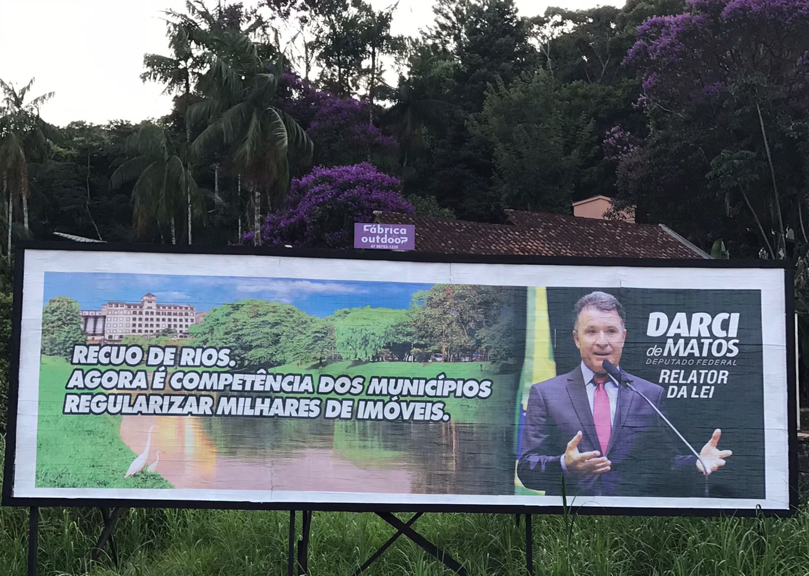 Outdoor mostra imagem ilustrativa de rio margeado por construções, com a foto do deputado Darci. A placa tem os seguintes dizeres "Recuo de rios. Agora é competência dos municípios regularizar  milhares de imóveis"