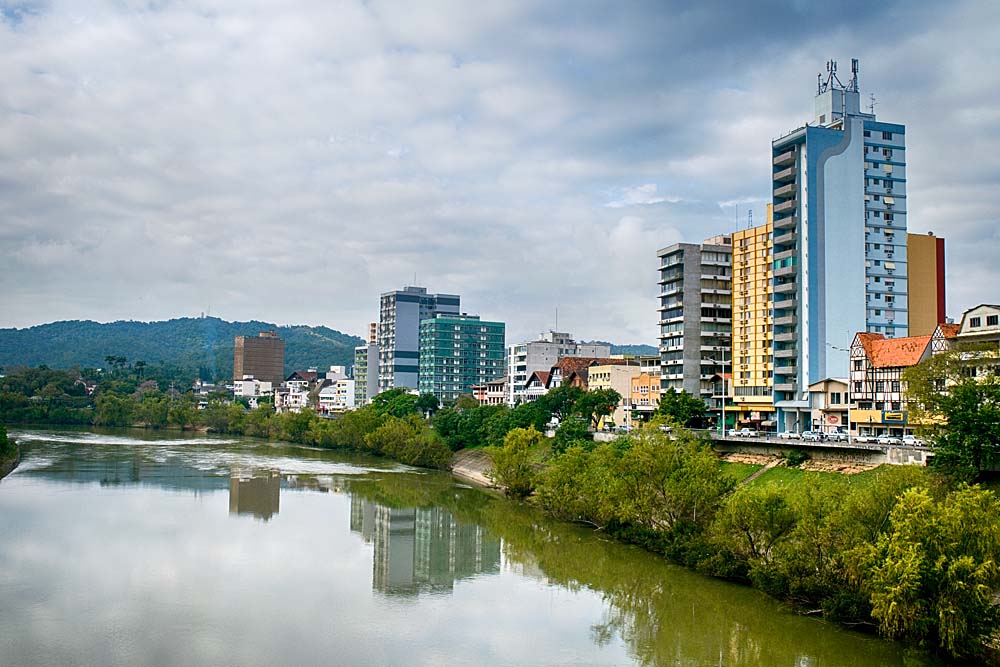 Trecho do rio Itajaí-Açu margeado por mata ciliar e construções como prédios e casas