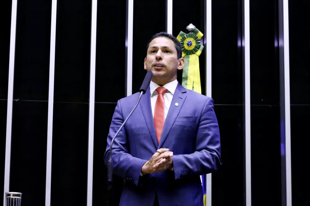 Marcelo Ramos é um homem branco com cabelos e olhos castanhos. Ele veste terno azul com gravata vermelha. Na imagem ele está discursando com um microfone à sua frente