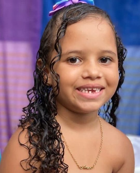 Heloysa era uma criança negra de cabelos castanhos e cacheados; na foto ela está vestindo um top roxo e uma saia verde, combinando com a decoração de festa de aniversário ao fundo; ela está sorrindo