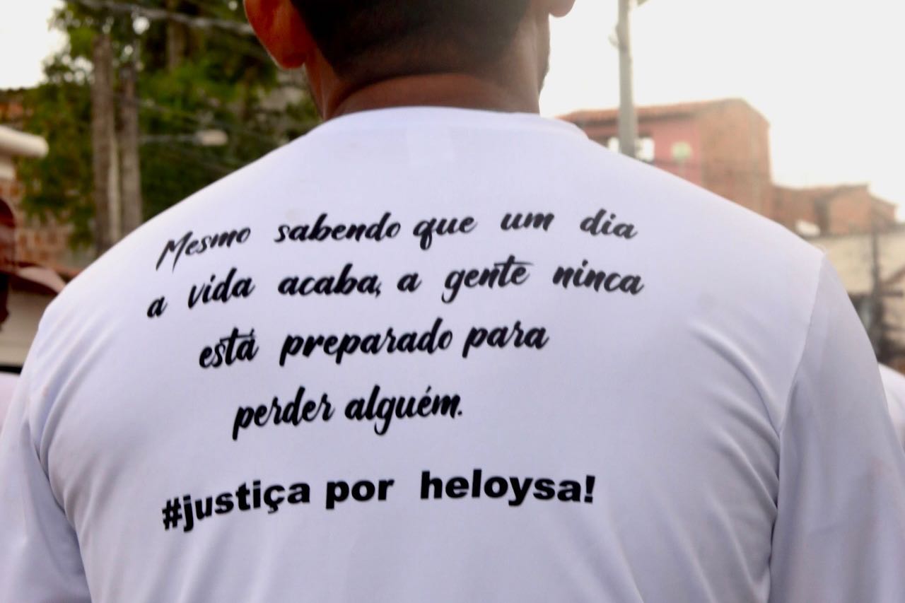 Imagem mostra as costas de um morador da comunidade, que veste camiseta branca com os dizeres "mesmo sabendo que um dia a vida acaba, a gente nunca está preparado para perder alguém. Justiça por Heloysa!"