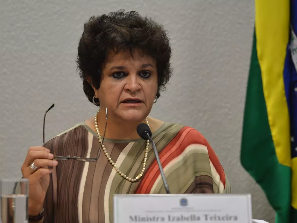 Ex-ministra do Meio Ambiente, Izabella Teixeira durante fala na Comissão Mista Permanente sobre Mudanças Climáticas; Teixeira é uma mulher branca com olhos e cabelos castanhos escuro; ela veste uma camiseta colorida em tons de marrom, branco e verde e segura o óculos na mão direita