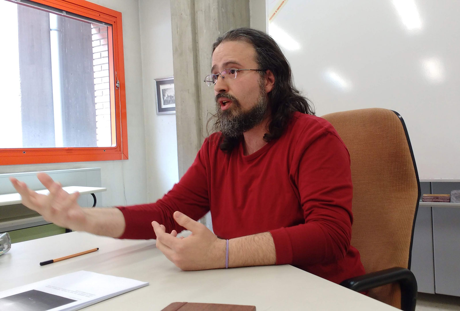 Fábio Candotti é um homem branco com barba e cabelo longo castanho; ele veste uma blusa de manga longa vermelha e está sentado em uma mesa branca