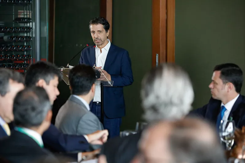 Joaquim Leite é um homem branco com cabelos castanhos, ele está vestindo um terno azul; Leite discursa para alguns convidados