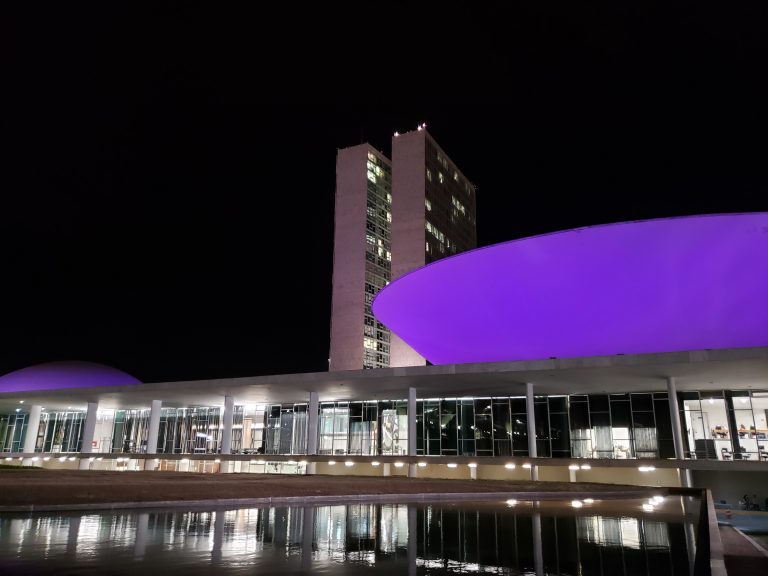 Prédio do Congresso iluminado de lilás, ao fundo a noite escura 