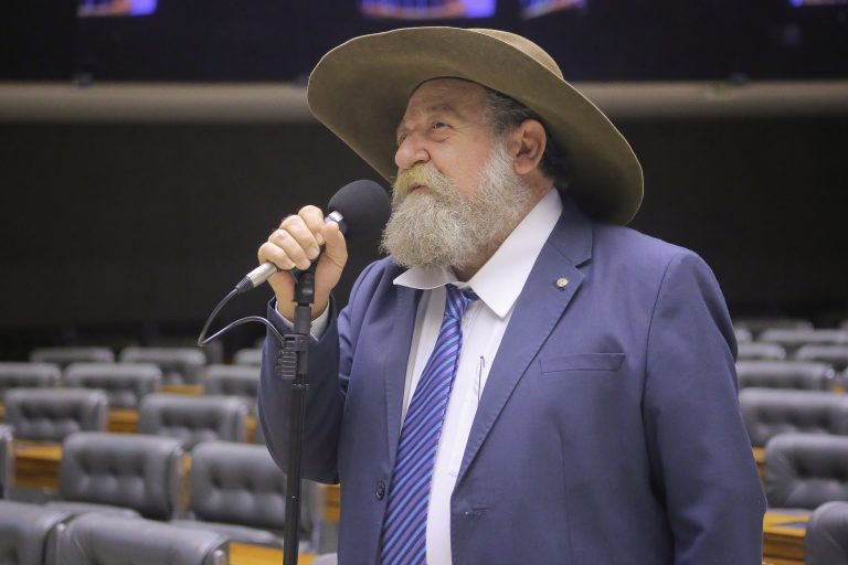 Deputado Nelson Barbudo fala ao microfone. Ele é branco, usa bigode e barba brancos e um chapéu 