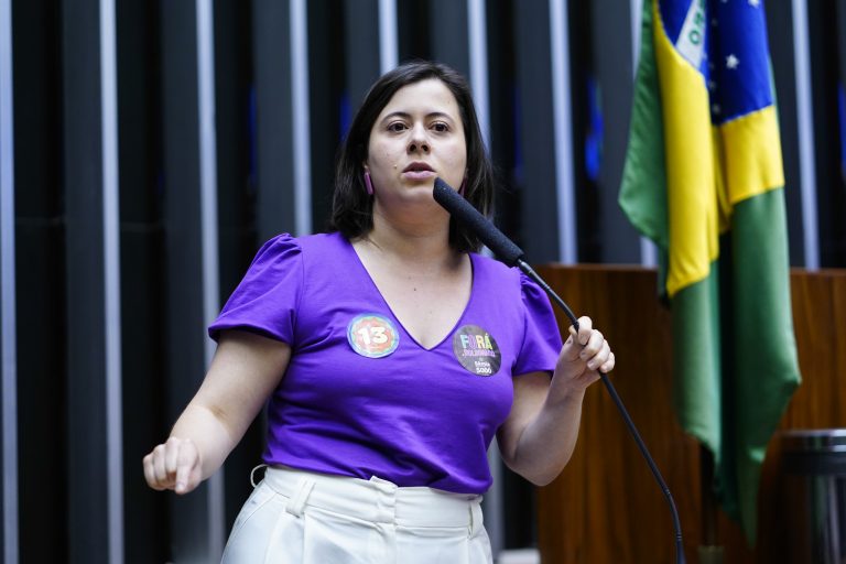 Deputada Sâmia Bomfim discursa no Plenário da Câmara. Ela é branca e usa uma blusa roxa. Ao fundo, à direita há uma bandeira do Brasil