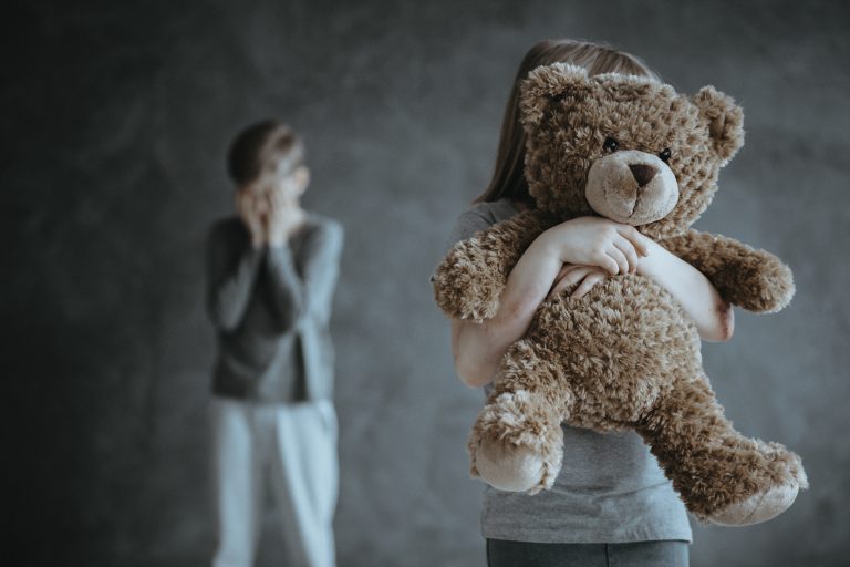 Uma menina segura um urso de pelúcia na frente do rosto dela. Ao fundo, desfocado, há um menino com as mãos no rosto