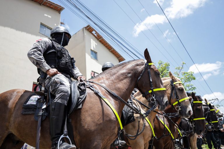 Um policial com o rosto coberto está sentado num cavalo. Atrás dele há vários cavalos enfileirados