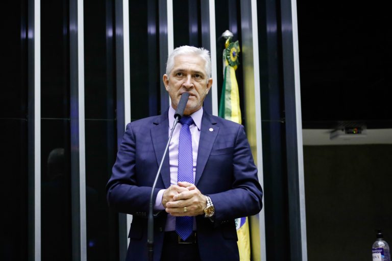 Deputado Milton Vieira fala ao microfone. Ele é um homem branco, com cabelo branco, usa um terno azul escuro, relógio e aliança