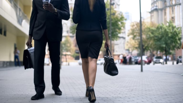 Um homem de terno preto caminha com um celular numa mão e uma pasta na outra. Ao lado, caminha uma mulher com um tailleur preto e uma bolsa na mão. A foto está cortada e não é possível ver os rostos